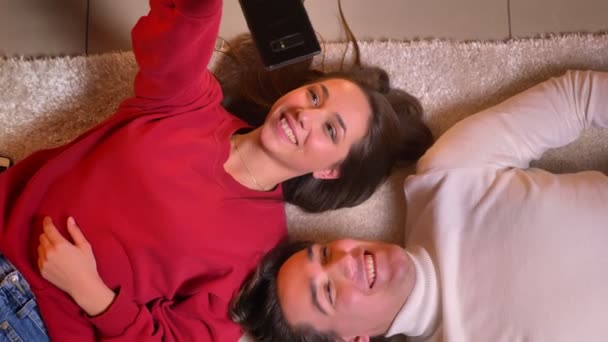 Top shot de joven hermosa pareja tumbada cabeza a cabeza en la alfombra y hacer selfies-fotos utilizando el teléfono inteligente en un ambiente acogedor . — Vídeo de stock