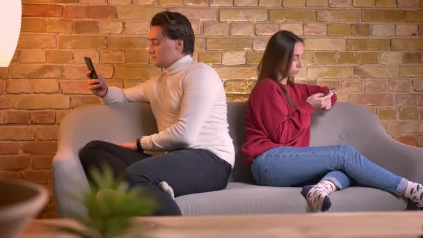 Portrait of young caucasian man and woman sit back to back on sofa with smartphone in home atmosphere . — Stok Video