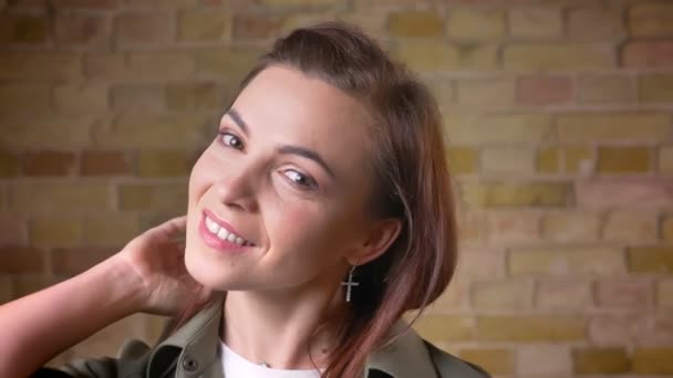 Portait af ung attraktiv brun-hovedet kvinde ryster hendes hår og smiler ind i kameraet på bricken væg baggrund . – Stock-video