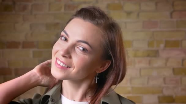 Portrait de jeune femme à la tête brune séduisante secouant les cheveux et souriant à la caméra sur fond de mur de poulet . — Video