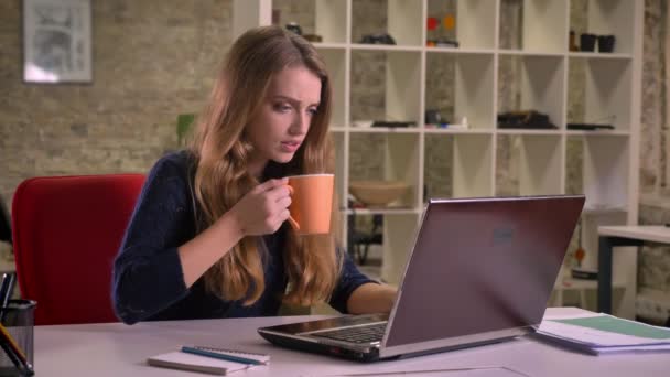 Porträt der hübschen blonden kaukasischen Geschäftsfrau, die im Büro mit Laptop arbeitet und Kaffee trinkt. — Stockvideo