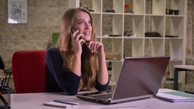 Sarışın beyaz iş kadını mutlu telefonda konuşurken office içinde laptop önünde portresi.