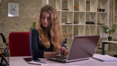 Portrait of busy blonde caucasian businesswoman working with laptop in office receives notification on tablet.