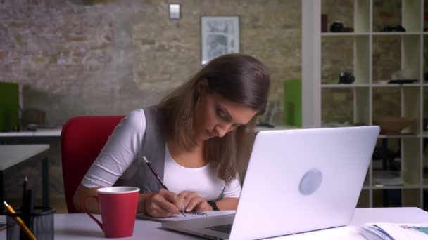 Attraktive Büroangestellte, die vor dem Laptop sitzt und sich Notizen macht und am Arbeitsplatz drinnen Kaffee trinkt — Stockvideo