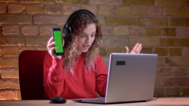 Portrét mladé ženy blogger v červená mikina ukazuje displej smartphone do notebooku na bricken zeď na pozadí. — Stock video