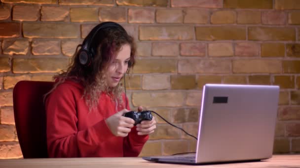 Porträtt av unga kvinnliga bloggare i röd Huvtröja spelar videospel med laptop och joystick på bricken vägg bakgrund. — Stockvideo