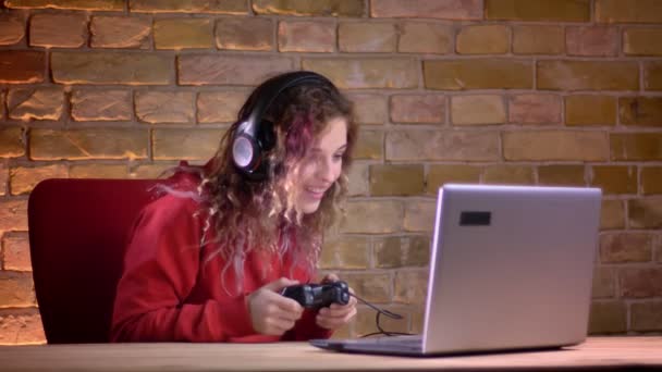 Porträt einer jungen Bloggerin in rotem Kapuzenpulli, die fröhlich Videospiel mit Joystick auf Backsteinmauern spielt. — Stockvideo