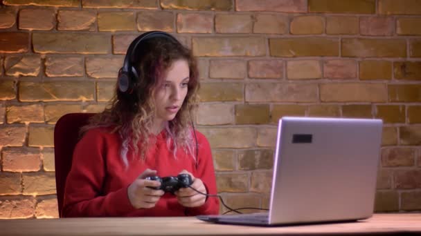 Portret van jonge vrouwelijke blogger in Rode hoodie afspelen van video game met behulp van de joystick en niet met schande op bricken muur achtergrond. — Stockvideo