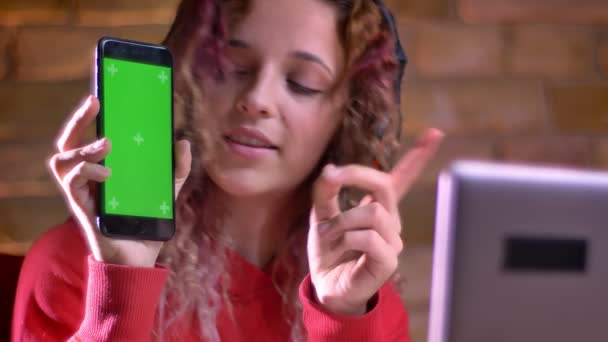 Portrét mladé ženy blogger v červená mikina ukazuje zelený displej chytrého telefonu do notebooku na bricken zeď na pozadí. — Stock video