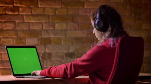 Vista trasera de cerca del joven blogger con capucha roja y auriculares que trabajan con computadora portátil sobre fondo de pared de ladrillo . — Vídeo de stock