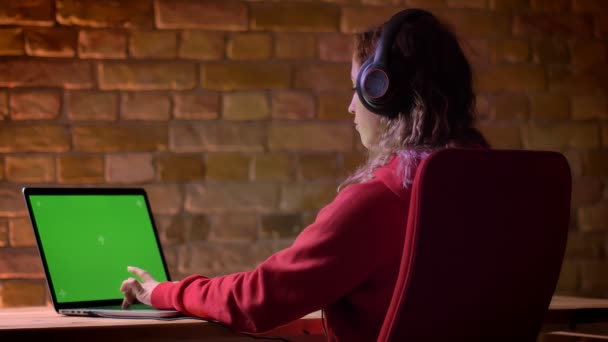 Visão traseira do jovem blogueiro com capuz vermelho e fones de ouvido trabalhando com laptop e falando no fundo da parede de tijolos . — Vídeo de Stock