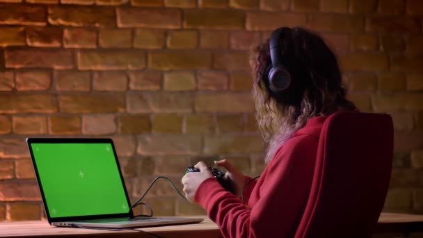 Vista trasera de un joven blogger con capucha roja y auriculares jugando videojuegos usando laptop y joystick sobre fondo de pared de ladrillo . — Vídeos de Stock