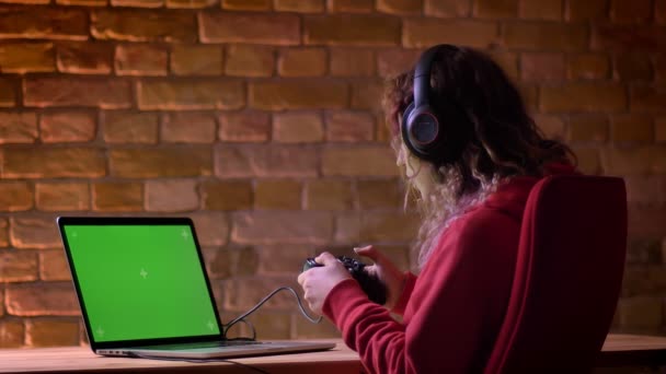 Vista trasera de un joven blogger con auriculares jugando felizmente a videojuegos usando laptop y joystick sobre fondo de pared de ladrillo . — Vídeos de Stock