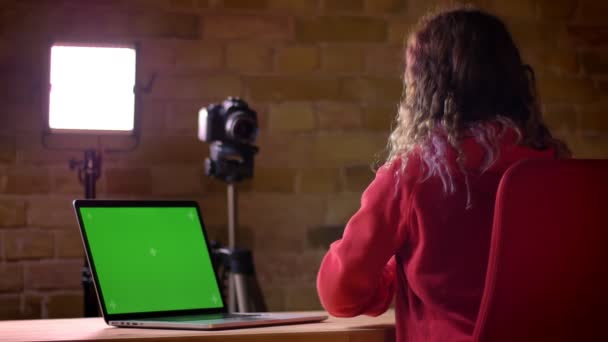 Vista trasera de un joven bloguero en auriculares grabando vídeo en cámara delante de un portátil sobre fondo de pared de ladrillo . — Vídeos de Stock
