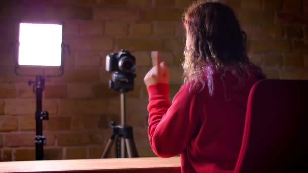 Vue arrière du jeune blogueur en sweat à capuche rouge enregistrant la vidéo devant la caméra et la lampe sur fond de mur de poulet . — Video