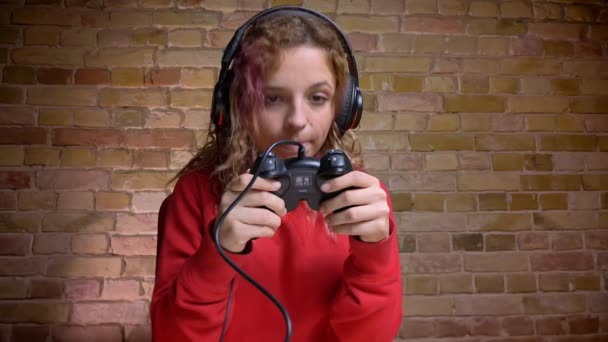 Close-up portret van attente vrouwelijke blogger in hoofdtelefoon met joystick voor camera op bricken muur achtergrond. — Stockvideo
