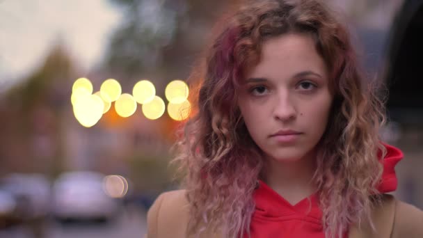 Primer plano retrato de la joven rubia caucásica mirando con calma en la cámara en el fondo borroso de la ciudad noche . — Vídeos de Stock