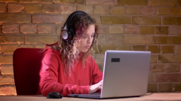 Porträtt av unga kvinnliga bloggare i röd Huvtröja gärna pratar med laptop på bricken vägg bakgrund. — Stockvideo