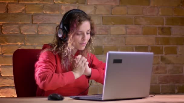 Retrato de jovem blogueira com capuz vermelho alegremente batendo palmas e falando usando laptop no fundo da parede de tijolos . — Vídeo de Stock