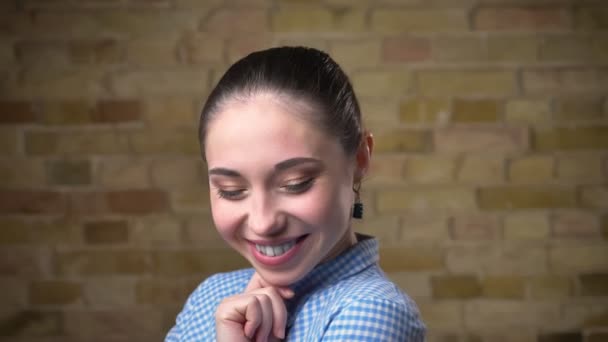 Gros plan portrait de femme brune caucasienne avec queue de cheval posant avec sourire sur la caméra sur fond de mur de poulet . — Video