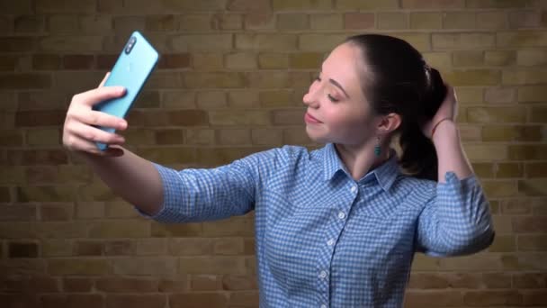 Portrait of caucasian woman with ponytail smilingly making selfie-photos using smartphone on bricken wall background. — Stock Video