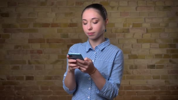 Portrait of caucasian woman with ponytail attentively watching into smartphone on bricken wall background. — Stock Video