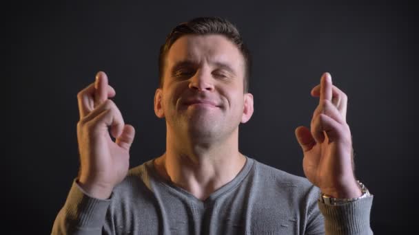 Retrato de hombre caucásico de mediana edad haciendo señas de dedos cruzados para mostrar esperanza en la cámara sobre fondo negro . — Vídeo de stock