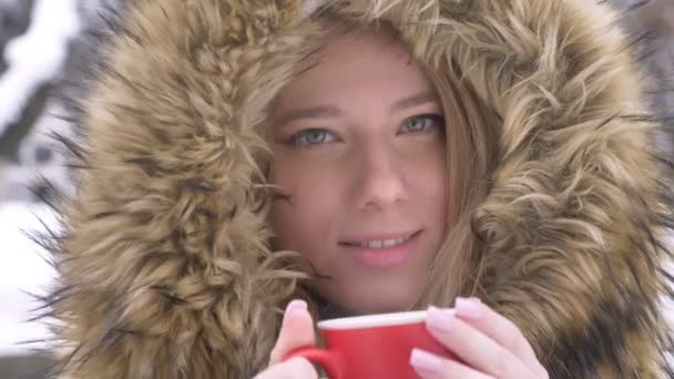 Close-up retrato de menina com xícara de bebida quente assistindo em câmera no fundo da rua de inverno . — Vídeo de Stock