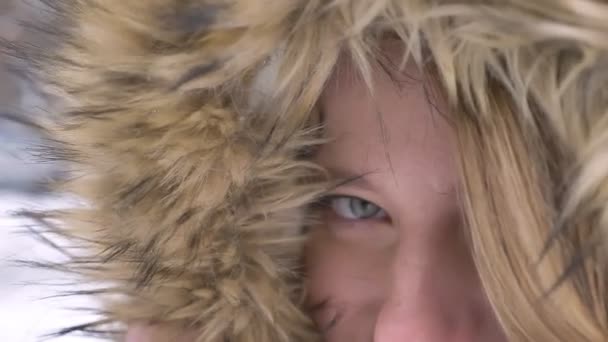 Nahaufnahme-Augenporträt der schönen jungen kaukasischen Mädchen mit Pelzhaube, die mit Flirt und Lächeln auf winterlichem Straßenhintergrund in die Kamera blickt. — Stockvideo