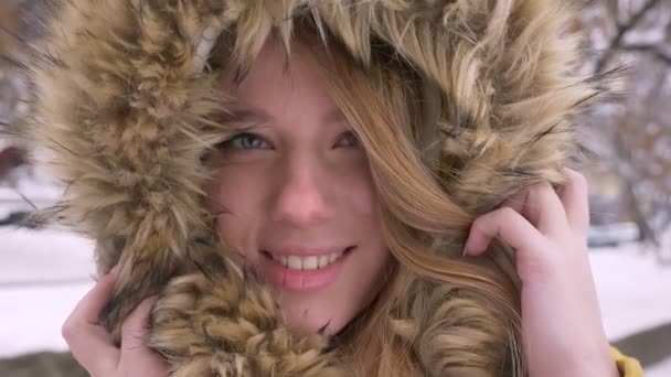 Close-up eye-portrait of beautiful young caucasian girl hiding in fur hood prettily smiling into camera on winter street background. — Stock Video