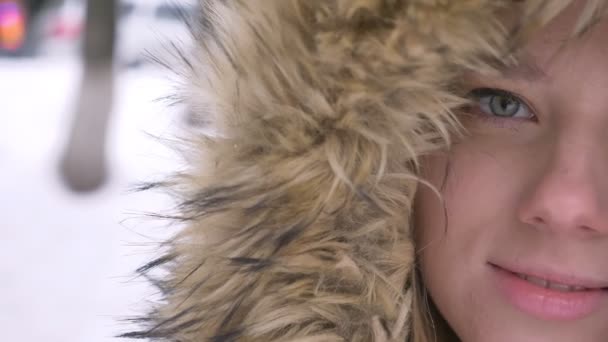 Close-up half-portrait of beautiful young caucasian girl in fur hood prettily smiling into camera on winter street background. — Stock Video