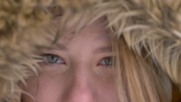 Close-up eye-portrait of attractive young caucasian girl in fur hood prettily smiling into camera on winter street background. — Stock Video