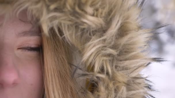 Close-up half-portrait of young caucasian girl in fur hood calmly watching into camera on winter street background. — Stock Video