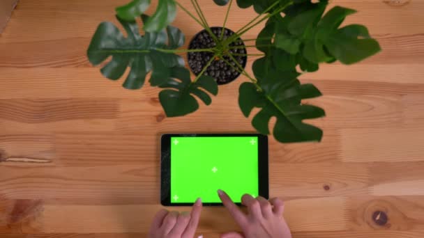 女性双手在木制桌子和植物背景上的水平平板电脑的绿色屏幕上打字的顶级镜头. — 图库视频影像