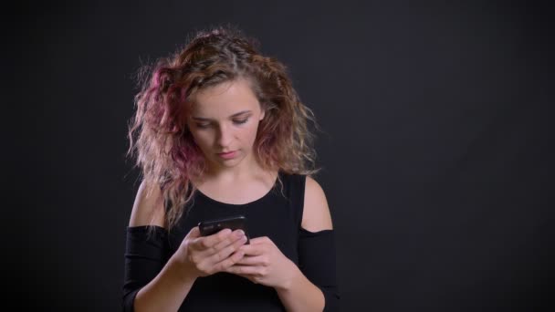 年轻的高加索女孩与粉红色的头发的肖像聚精会神地看着她的智能手机在黑色背景 — 图库视频影像