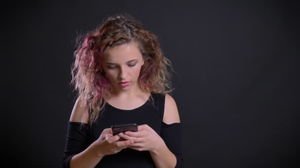 Retrato de jovem caucasiana com cabelo rosa assistindo em seu smartphone fica severamente divertido no fundo preto . — Vídeo de Stock