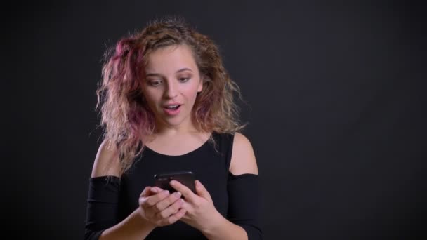 Ritratto di giovane ragazza caucasica con i capelli rosa guardando nel suo smartphone ottiene sorpreso e molto felice su sfondo nero . — Video Stock