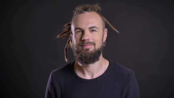 Close-up portrait of middle-aged caucasian man with dreadlocks and piercing watching smilingly into camera on black background. — Stock Video