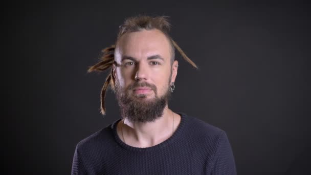 Portrait of caucasian man with dreadlocks and piercing watching calmly into camera and showing his teeth on black background. — Stock Video