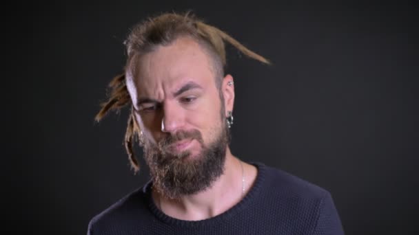 Portret van charismatische man met dreadlocks en piercing serieus en zorgvuldig in de camera kijken op zwarte achtergrond. — Stockvideo