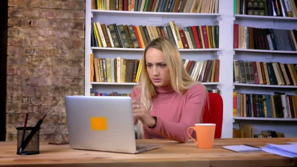 Портрет панічної білої дівчини з пірсингом, що працює з ноутбуком і стикається з проблемами на фоні книжкових полиць . — стокове відео