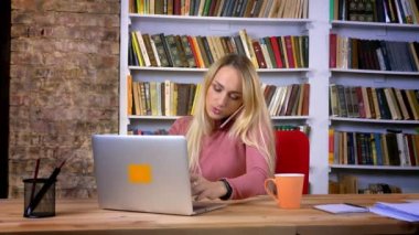 Dikkatle laptop ile çalışma ve kitap telefonda konuşurken piercing ile genç beyaz kız portresi arka plan raflar