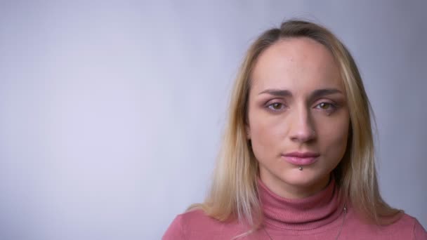 Retrato de atractiva mujer rubia caucásica mirando en serio y parpadeando en la cámara sobre fondo gris . — Vídeos de Stock