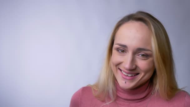 Porträt einer jungen blonden Kaukasierin, die schüchtern in die Kamera lacht, auf grauem Hintergrund. — Stockvideo