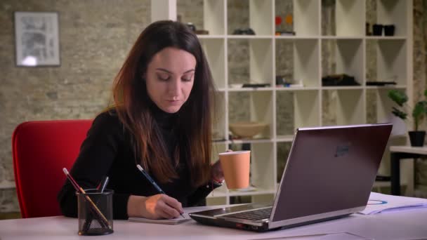 Porträt einer jungen brünetten Geschäftsfrau, die Notizen ins Kopierbuch macht und im Büro Kaffee trinkt. — Stockvideo