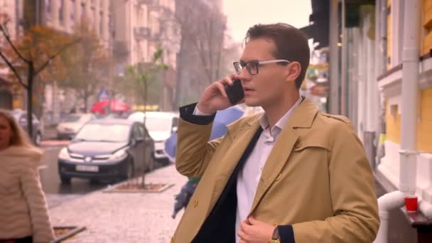 在一个秋日, 有吸引力的休闲白种人在电话里交谈, 站在城市路过的汽车旁的人行道上 — 图库视频影像