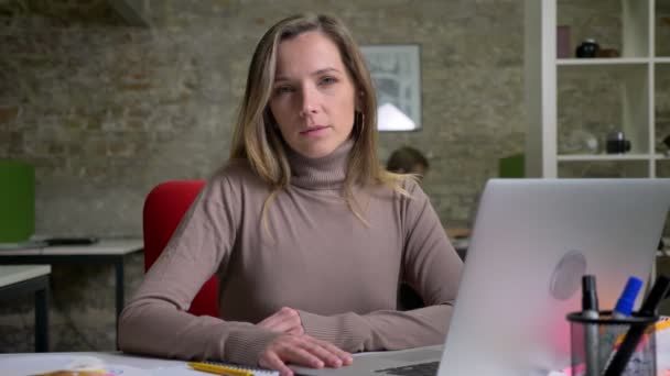 Primer plano retrato de atractiva mujer trabajadora de oficina caucásica en frente de la computadora portátil mirando a la cámara y sonriendo — Vídeo de stock