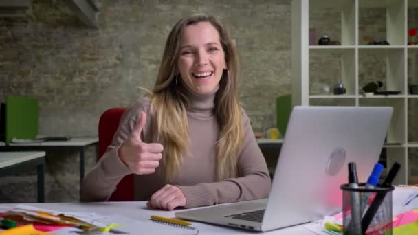 Портрет крупным планом симпатичной офисной работницы, сидящей перед ноутбуком, улыбающейся и показывающей большой палец вверх — стоковое видео