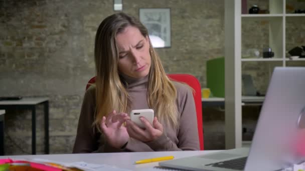 Closeup-portrett av moderne kvinnelige ansatte som leser i telefonen og ler sittende ved siden av laptopen på arbeidsplassen – stockvideo