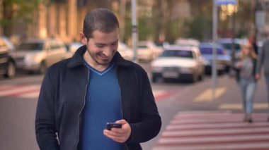 Çapraz-yürüyüş üzerinde duran ve arka plan üzerinde bulanık yürüyen insanlarla smartphone izlerken beyaz adam portresi.